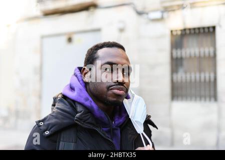 Afroamerikanischer Rüde in Kapuzenpullover und Oberbekleidung, der die Gesichtsmaske auszieht und auf den verschwommenen Hintergrund des Gebäudes auf der Stadtstraße blickt Stockfoto