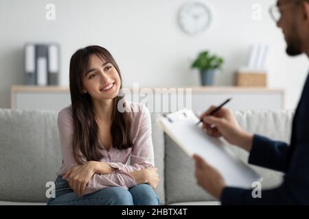 Junge arabische Klientin, die mit Psychologin spricht und lächelt, auf der Couch in der Klinik sitzt Stockfoto