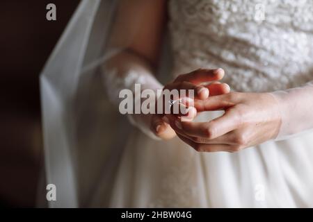 Zugeschnittenes Foto von sanften Händen der Braut, die in einem weißen Spitzenkleid steht, setzt einen Ehering auf ihren Finger. Hochzeitstag. Stockfoto