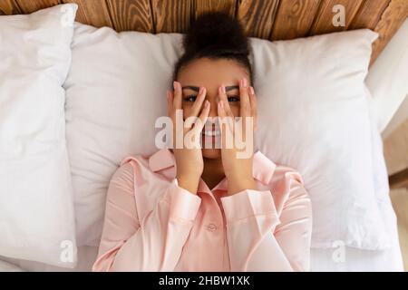 Guten Morgen. Draufsicht Porträt einer hübschen afroamerikanischen Dame, die im Bett liegt, durch die Finger schaut und lächelt Stockfoto