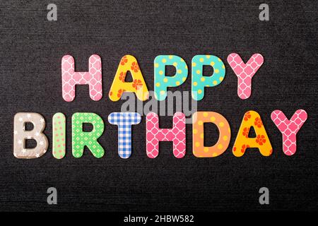 Karte mit Happy Birthday-Wörtern aus gemischten, lebendigen farbigen Holzbuchstaben auf einem strukturierten, dunkelschwarzen Textilmaterial, das als Botschaft verwendet werden kann Stockfoto