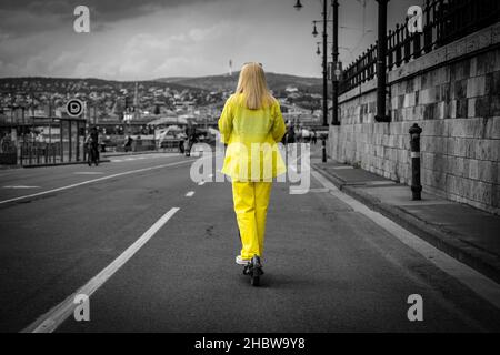 Blonde Frau in gelben Kleidern mit einem Elektroroller auf dem Donauwrack in Budapest, von hinten geschossen, gelb auf schwarz-weißem Hintergrund Stockfoto