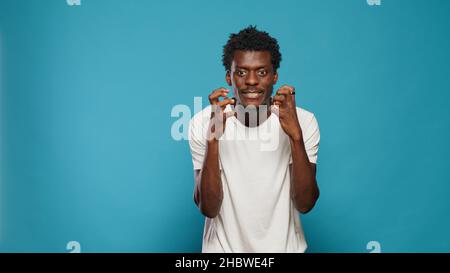 Frustrierter Erwachsener, der sich im Studio wütend fühlt und sich mit den Fäusten festhält. Unzufriedener Mensch, der wegen Wut und Frustration Zähne zeigt und auf blauem Hintergrund steht. Porträt eines aggressiven Mannes Stockfoto