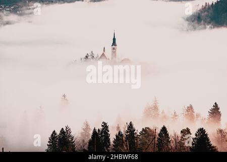 Erstaunliches Panorama des Bleder Sees Blejsko Jezero an einem nebligen Morgen mit der Wallfahrtskirche Mariä Himmelfahrt auf einer kleinen Insel und dem Schloss Bled Stockfoto