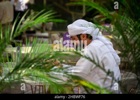 Dubai, VAE - 11.30.2021: Arabischer Mann mit Bart in traditionellem weißen Kleid (taub) sitzt hinter Palmblättern, während er sein Mobiltelefon in Saudi-Arabien benutzt Stockfoto