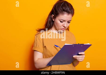 Junge Frau, die sich im Studio Notizen zu Lehrbuchpapieren mit Stift gemacht hat. Konzentrierte Person, die auf Notizbuchdateien schreibt und Dokumente und Unterlagen über das Projekt auf der Notizblock-Zwischenablage unterschreibt. Stockfoto