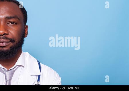 Nahaufnahme mit Halbgesicht Porträt eines lächelnden Arztes in weißem Laborkittel mit Stethoskop auf blauem Hintergrund. Beschnittenes Bild eines afroamerikanischen Mediziners, der selbstbewusst und entspannt aussieht. Stockfoto