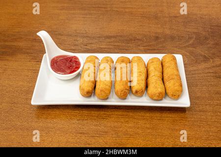 Frisch zubereitete venezolanische tequeños auf einem weißen rechteckigen Teller, Erdbeersirup und Holztisch Stockfoto