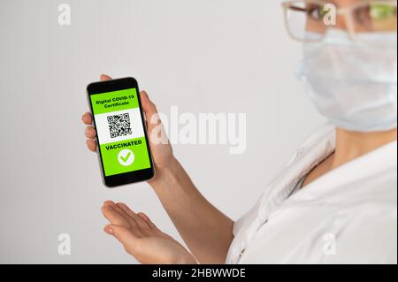 Der Arzt empfiehlt eine Impfung und hält ein Smartphone mit einem QR-Code in der Kamera. Stockfoto