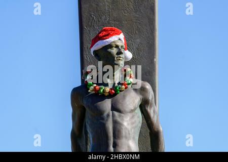 Surfer-Statue, Surfdenkmal, Bronze eines männlichen Surfers, der vor seinem mit Weihnachtsmütze und Weihnachtsleis geschmückten Brett steht für Christma Stockfoto