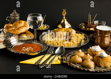 Ramadan-Tisch mit Tomatensuppe, Hühnerhühnern auf Reis, Ramadan-Brot und türkischem Dessert auf der schwarzen Oberfläche. Gebrauchte Küchengeräte aus Kupfer und Silber Stockfoto