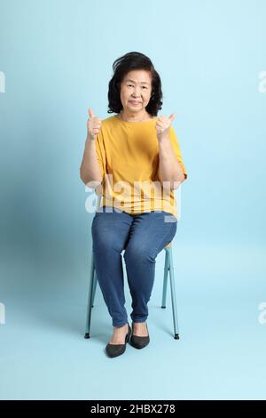 Die ältere asiatische Frau sitzt auf dem blauen Hintergrund. Stockfoto
