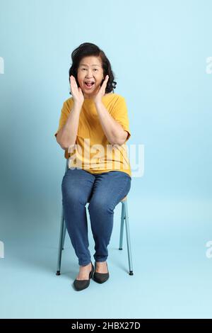 Die ältere asiatische Frau sitzt auf dem blauen Hintergrund. Stockfoto