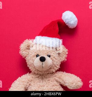 Weihnachtsgeschenk. Teddybär trägt Weihnachtsmütze auf rotem Hintergrund, Weihnachtsgeschenke, Spielzeug und Stofftiere. Weihnachtskarte. Stockfoto