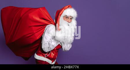 Der schlinkende weihnachtsmann trägt eine riesige Geschenktüte auf violettem Hintergrund, während er die Kamera anschaut. Stockfoto