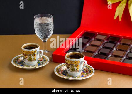 Traditioneller türkischer Kaffee in vergoldeten Tassen, auf der braunen Oberfläche mit madlen-Schokolade und einem Glas Wasser. Stockfoto