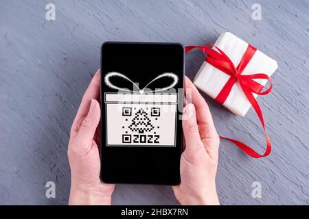 Hände, die ein Mobiltelefon mit einem schwarzen Bildschirm mit einer gezeichneten Geschenkbox und einem qr-Code in Form eines Weihnachtsbaums und einer Geschenkbox mit einem roten Band halten Stockfoto