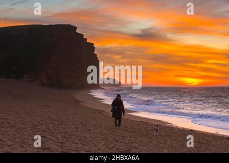 West Bay, Dorset, Großbritannien. 22nd. Dezember 2021. Wetter in Großbritannien. Ein Hundewanderer, der bei Sonnenaufgang einen Spaziergang Unternehmen kann, während die Wolken über dem Strand und den Klippen der West Bay an der Dorset Jurassic Coast orange leuchten. Bildnachweis: Graham Hunt/Alamy Live News Stockfoto