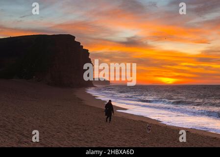 West Bay, Dorset, Großbritannien. 22nd. Dezember 2021. Wetter in Großbritannien. Ein Hundewanderer, der bei Sonnenaufgang einen Spaziergang Unternehmen kann, während die Wolken über dem Strand und den Klippen der West Bay an der Dorset Jurassic Coast orange leuchten. Bildnachweis: Graham Hunt/Alamy Live News Stockfoto