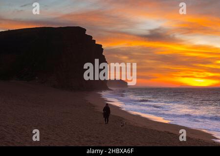 West Bay, Dorset, Großbritannien. 22nd. Dezember 2021. Wetter in Großbritannien. Ein Hundewanderer, der bei Sonnenaufgang einen Spaziergang Unternehmen kann, während die Wolken über dem Strand und den Klippen der West Bay an der Dorset Jurassic Coast orange leuchten. Bildnachweis: Graham Hunt/Alamy Live News Stockfoto