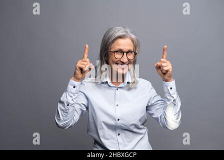 Lächelnde ältere grauhaarige Geschäftsfrau, isoliert auf grauem Hintergrund, zeigt den Finger nach oben. Dame, die auf etwas achtet Stockfoto