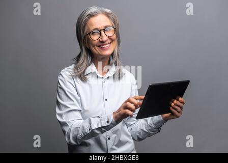 Moderne ältere ältere Frau in formeller Kleidung mit einem digitalen Tablet isoliert auf grau. Porträt einer reifen weiblichen Büroangestellerin, die Online-Technologie für Geschäftsabläufe nutzt, Computer-App für die Buchhaltung Stockfoto