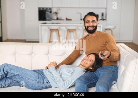 Horizontale Aufnahme des verliebten indischen Paares, das auf dem Sofa sitzt und mit Vergnügen lächelt, vor dem Fernseher schaut. Multirassischer Mann, der die Fernbedienung zur Hand hält. Home-Konzept Stockfoto