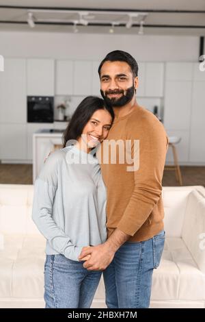 Lächelnder Mann umarmt seine charmante Frau. Zwei Menschen stehen und blicken freudig auf die Kamera. Das junge indische Paar verbringt gerne Zeit in einer gemütlichen modernen Küche zu Hause Stockfoto