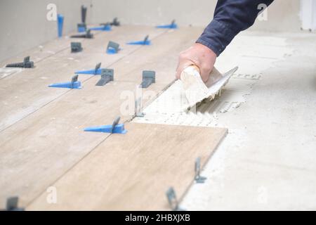 Nicht erkennbarer Maurer, der Fliesen mit einer gekerbten Kelle auf den Boden klebte. Stockfoto