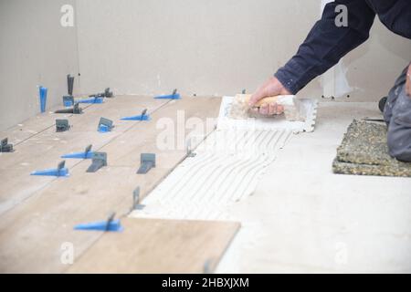 Arbeiter reicht die Hände, die Fliesen mit der eingekerbten Kelle auf den Boden kleben. Stockfoto