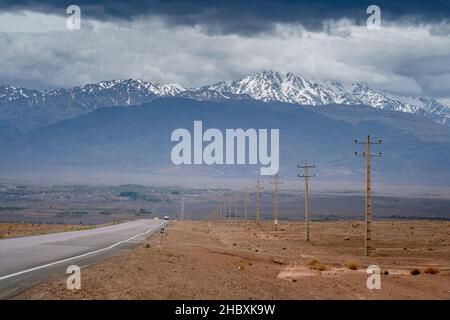Komprimierte Aufnahme einer langen geraden Rayen-Straße mit elektrischen Polen entlang, die zur Stadt Rayen in der Provinz Kerman, Iran, führt. Majestätischer Hazaran- oder Hezar-Gipfel Stockfoto