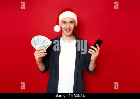 Aufgeregt Mann mit Geld und Smartphone in den Händen gekleidet Weihnachtsmann Weihnachtsmütze freut sich zu gewinnen Stockfoto