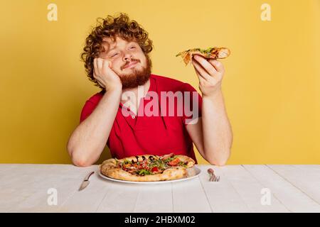 Porträt eines rotbärtigen Mannes, der köstliche italienische Pizza auf gelbem Studiohintergrund verkostet. Stockfoto