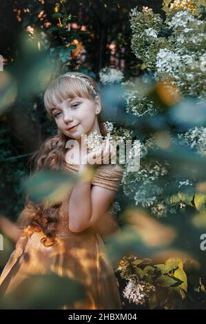 Schöne 11-jährige Mädchen mit langen blonden Haaren in einem Dickicht von Hortensien. Das Bild einer charmanten kleinen Prinzessin. Mädchen schaut in die Linse. Stockfoto