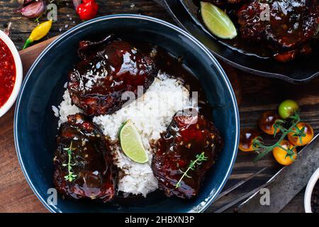 Draufsicht auf ein würziges Chili Chicken Oberschenkelfleisch serviert mit weißem Reis über einem Holzschneidebrett und rustikalem Hintergrund mit frischen Zutaten von heißem SA Stockfoto
