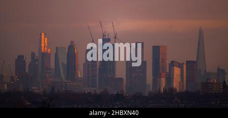 London, Großbritannien. 22. Dezember 2021. Wechselnde Lichtverhältnisse auf Londoner Wolkenkratzern während eines farbenfrohen Wintersonnenaufgangs, bei dem Sonnenlicht die oberen Stockwerke der höchsten Bürogebäude trifft. Aufsteigendes Sonnenlicht trifft auf die Spitze von 22 Bishopsgate (3rd links im Bild), dem höchsten Gebäude in der City of London. Die Entwicklung von Hochhauswohnungen in Vauxhall (Mitte im Bild mit Baukräne) verdeckt allmählich einige der Bürogebäude der Stadt in der „Square Mile“ von Wimbledon aus gesehen. Kredit: Malcolm Park/Alamy Stockfoto