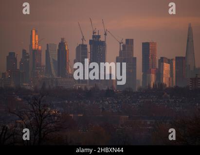 London, Großbritannien. 22. Dezember 2021. Wechselnde Lichtverhältnisse auf Londoner Wolkenkratzern während eines farbenfrohen Wintersonnenaufgangs, bei dem Sonnenlicht die oberen Stockwerke der höchsten Bürogebäude trifft. Aufsteigendes Sonnenlicht trifft auf die Spitze von 22 Bishopsgate (3rd links im Bild), dem höchsten Gebäude in der City of London. Die Entwicklung von Hochhauswohnungen in Vauxhall (Mitte im Bild mit Baukräne) verdeckt allmählich einige der Bürogebäude der Stadt in der „Square Mile“ von Wimbledon aus gesehen. Kredit: Malcolm Park/Alamy Stockfoto