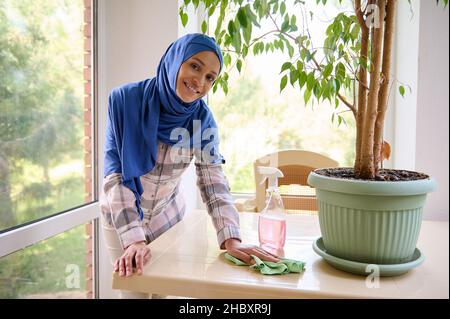 Entzückende wunderschöne, charmante arabische Muslimin mittleren Alters mit einem bedeckten Kopf in blauem Hijab, der während des Frühlings Staub auf dem Tisch auf der Veranda abwischt Stockfoto