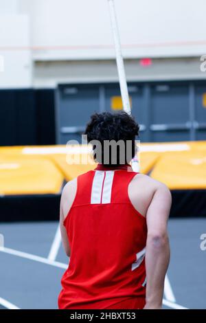 Ein High-School-Athlet auf dem Laufsteg mit einem Stock in den Händen und den Matten im Hintergrund bereit, bei einem Indoor-Wettbewerb Stabhochsprung zu machen. Stockfoto