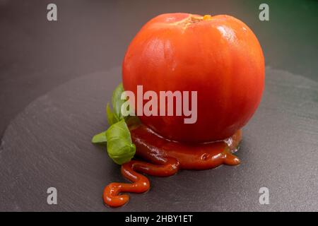 Geschälte Tomate mit Ketchup und Basilikumblättern, isoliert auf einer schwarzen Steinfliese Stockfoto