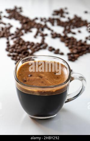 Transparente Tasse Kaffee mit viel Crema und verschwommenen Kaffeebohnen im Hintergrund Stockfoto