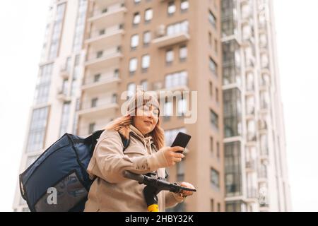 Low-Angle-Ansicht der Lebensmittelzustellung Frau Kurierin mit Thermobeutel und Elektroroller, in der Stadt verloren und mit Navigation im Telefon Stockfoto