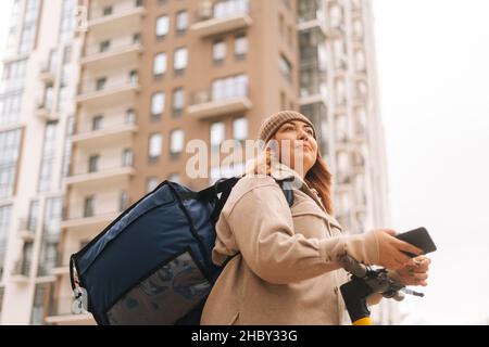 Nahaufnahme aus dem unteren Winkel der Frau, die Lebensmittel geliefert hat, mit Thermo-Rucksack, der das Mobiltelefon hält, das auf die nächste Bestellung wartet und mit einem Elektroroller steht. Stockfoto