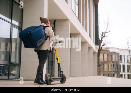 Low-Winkel-Rückansicht von unkenntlich weiblichen Kurier Lebensmittel Lieferung mit großen thermischen Rucksack zu Fuß mit Elektroroller in der Stadt Straße. Stockfoto