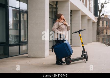 Weibliche Kurierdienst Lebensmittelzustellung mit Thermorucksack suchen Client nach Gespräch auf Handy stehen mit Elektroroller in der Straße. Stockfoto