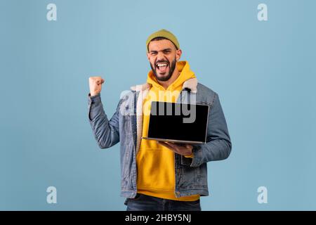 Ja. Glücklicher arabischer Mann hält pc-Laptop mit leerem Bildschirm schütteln geballte Fäuste machen Sieger Geste Stockfoto
