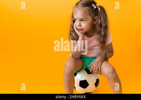 Authentische niedliche lächelnde Vorschule kleines Mädchen mit klassischen schwarz-weißen Fußball Blick auf die Kamera auf gelbem Hintergrund. Kind spielen Fußball in t Stockfoto