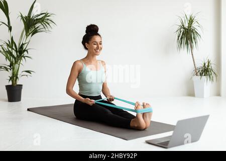 Lächelnde Frau trainiert mit elastischem Widerstandsgummi vor dem Laptop Stockfoto
