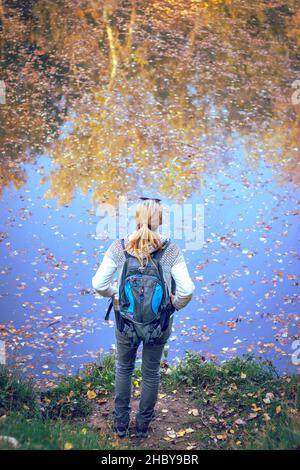 Wanderfrau, die in der Herbstsaison am See steht, Spiegelung der Bäume auf der Wasseroberfläche Stockfoto
