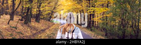 Frau mit Strickmütze und Rucksack steht in der Herbstsaison mitten im Wald auf der Straße, Panoramabild. Frau und ihre Reise. Stockfoto